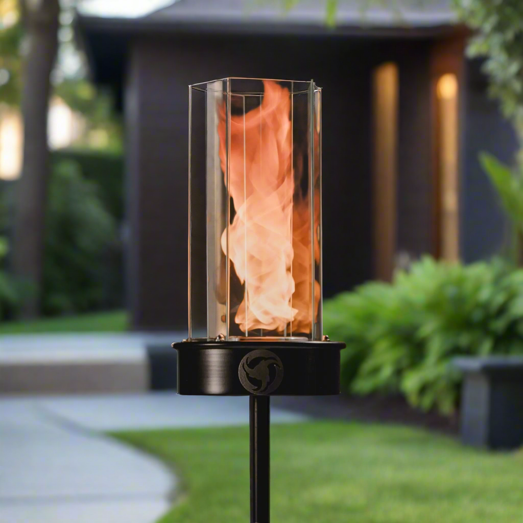Outdoor oil tiki torch with a tall vertical flame, clear glass wind guard, and black metal post mounted on a ground stake, designed for modern patio, garden, and landscape lighting.