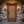 Outdoor oil wall-mount fire feature with a vertical flame enclosed in a clear glass wind guard, supported by a curved metal arm and wall plate, designed for modern patio, garden, and exterior wall lighting.