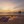 A beach scene at sunset with a small black fire pit containing a flame in the foreground, a colorful striped towel on the sand, and two silhouetted people walking near the shoreline in the distance.