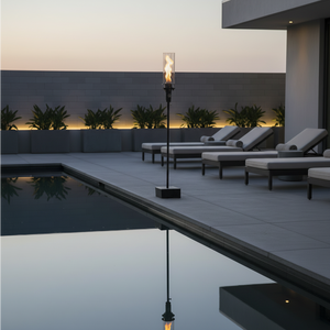Outdoor gas tiki torch featuring a tall vertical flame, clear glass wind guard, and black metal base for modern patio, garden, and landscape lighting using propane or natural gas.