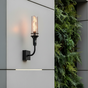 Outdoor wall-mounted gas tiki torch with a tall vertical flame, clear glass wind guard, black metal wall mount arm, and rectangular wall plate, designed for modern patio, garden, and exterior wall lighting using propane or natural gas.