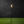 A single black outdoor torch light with a clear glass cylinder and visible orange-yellow flame, standing on bright green artificial grass against a dark charcoal background.