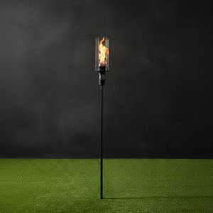 A single black outdoor torch light with a clear glass cylinder and visible orange-yellow flame, standing on bright green artificial grass against a dark charcoal background.
