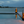 Outdoor patio scene at dusk with a rectangular pool, stone tile deck, two black metal fire pits with bright orange flames, and a waterfront view across calm water under a cloudy blue sky.