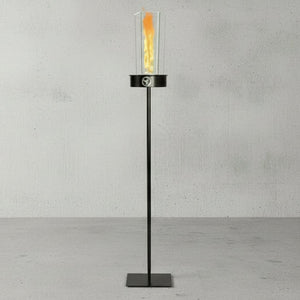 Outdoor oil tiki torch with a tall vertical flame, clear glass wind guard, and metal pole attached to an in-ground stake, designed for modern patio, garden, and landscape lighting.