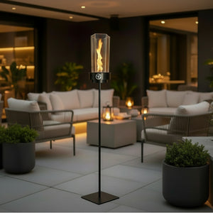 A tall outdoor patio fire pillar with a clear glass cylinder and visible flame, mounted on a slim black metal stand with a square base, shown in a modern patio lounge setting at night.