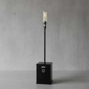 Black outdoor lamp with a cylindrical shade on a concrete floor against a gray wall.