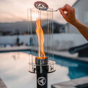Hand holding a portable fire pit with flames by a poolside at sunset.