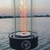 Outdoor oil tabletop fire feature with a vertical flame enclosed in a clear glass wind guard, designed for modern patio tables, outdoor dining areas, and ambient lighting.