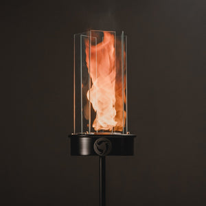 Outdoor oil tiki torch with a tall vertical flame, clear glass wind guard, and black metal post mounted on a ground stake, designed for modern patio, garden, and landscape lighting.