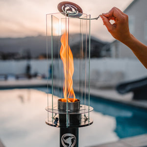 A Tiki torch with a flame, featuring a metal pole and glass enclosure, placed on a pool deck.